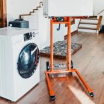 SuperHandy GUO097 material lift holding an appliance raised on its forks next to a washing machine